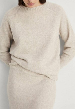 Pull en laine beige avec un col rond, des poignets côtelés et un ourlet. Le tissu a un aspect doux et texturé. Associé à une jupe assortie.
