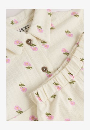 Kremowy materiał sukienki ozdobiony haftem z różowymi kwiatami i zielonymi liśćmi. Posiada kołnierzyk i marszczone detale falbanek.