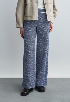 Femme portant un pantalon large en tweed bleu, une veste courte beige, un haut blanc et des mocassins marron foncé, debout sur un sol gris.