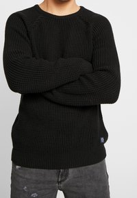 Homme portant un pull en tricot noir texturé, les bras croisés sur la poitrine, associé à un jean noir délavé et usé.