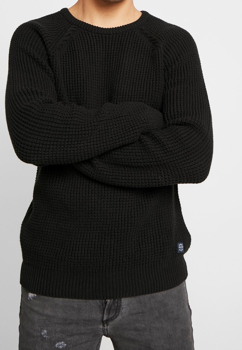 Homme portant un pull en tricot noir texturé, les bras croisés sur la poitrine, associé à un jean noir délavé et usé.