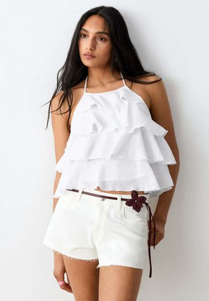 Jeune femme portant un haut dos nu blanc à volants et un short en jean blanc avec ceinture marron foncé et détail de fleur, debout devant un mur blanc.