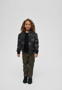 Veste camouflage noire et grise avec une fermeture éclair, t-shirt noir, pantalon cargo vert olive et chaussures noires, sur un enfant avec des cheveux bouclés.