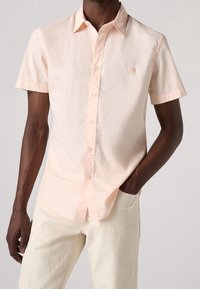 Camiseta de manga corta color rosa claro hecha de tela suave, con diseño de botones, cuello y un logo sutil en el pecho. Combinada con pantalones color crema.