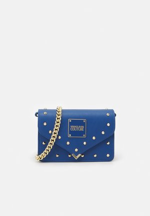 Niebieska torba typu kopertówka z ćwiekami, złotym łańcuszkowym paskiem i plakietką z logo Versace Jeans Couture na przedniej klapie.