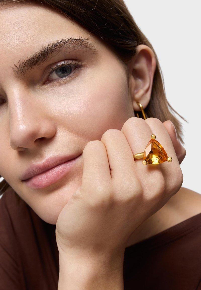 Złoty pierścień z trójkątnym bursztynowym kamieniem i trzema kulistymi akcentami; gładka faktura; noszony na dłoni na neutralnym tle.