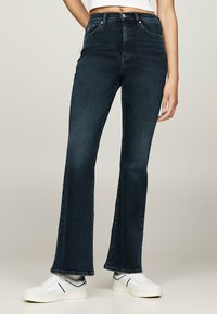 Jeans de tiro alto acampanados en denim azul oscuro con una textura suave y un desvanecimiento mínimo, combinados con zapatillas blancas con acentos azules.
