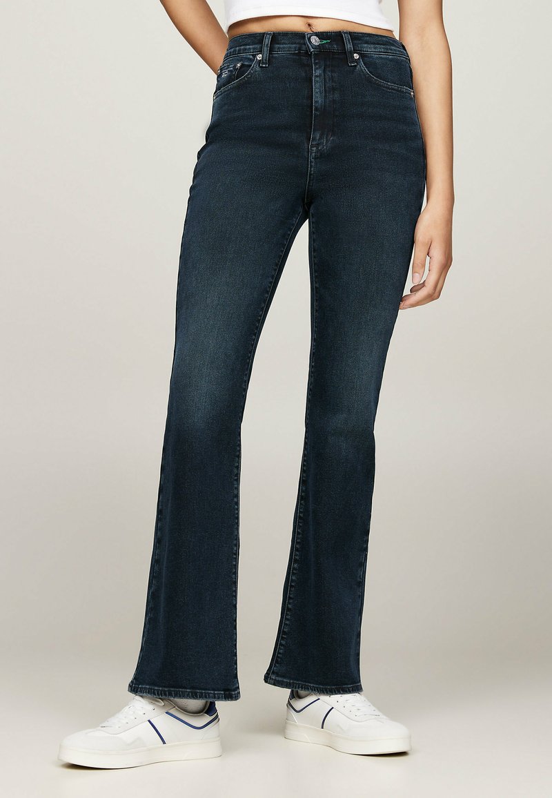Jeans de tiro alto acampanados en denim azul oscuro con una textura suave y un desvanecimiento mínimo, combinados con zapatillas blancas con acentos azules.