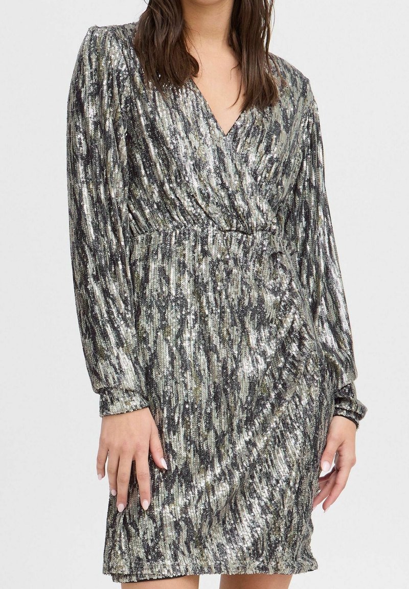 Femme portant une robe argentée à sequins avec manches longues, col en V, longueur genou, comportant des motifs noirs et gris, debout devant un fond uni.