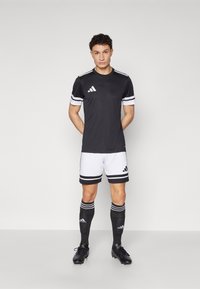 Maillot de sport noir avec des rayures blanches, shorts noirs avec des accents blancs, chaussettes noires et chaussures de football noires. Marque Adidas visible.