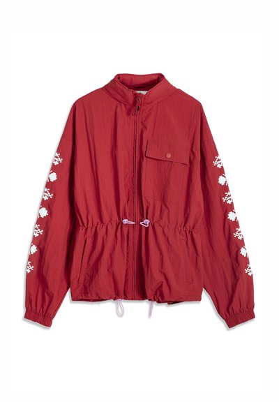 Veste rouge légère avec fermeture éclair frontale, poche poitrine à bouton-pression, poignets élastiques, motif pixellisé blanc sur les manches, et cordons ajustables à la taille.