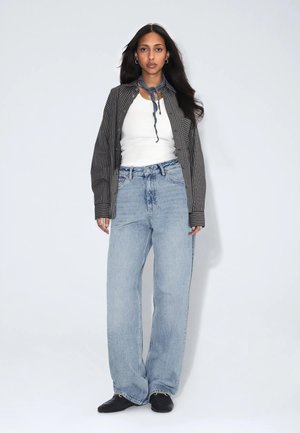 Kvinde iført en ternet skjorte over en hvid tank top, blå viddebenede jeans, sorte sko og et lille blåt tørklæde bundet omkring halsen.