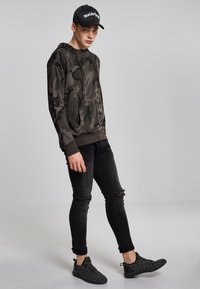 Kamuflasjemønstret hettegenser i mørkegrønn og svart, kombinert med slitte svarte skinny jeans og teksturerte svarte joggesko. Svart caps med logo.