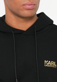 Primer plano de una persona que lleva una sudadera negra con capucha con el logo dorado de KARL LAGERFELD en el pecho y cordones negros.