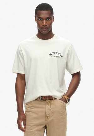 Camiseta de algodón color crema con mangas cortas que presenta un gráfico de "SUPERDRY NEW YORK" en negro. Combinada con pantalones caqui y un cinturón marrón.