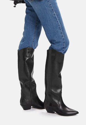 Černé kožené kozačky sahající po kolena s špičatou špičkou a nízkým, zdobeným podpatkem. Hladká textura s detaily bočního šití. Stylizováno s modrými džíny.