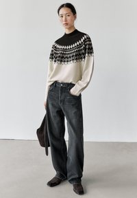 Femme portant un pull à motifs noir et blanc, un jean noir ample, des chaussures sombres à bout carré, tenant un sac foncé dans une pièce blanche.