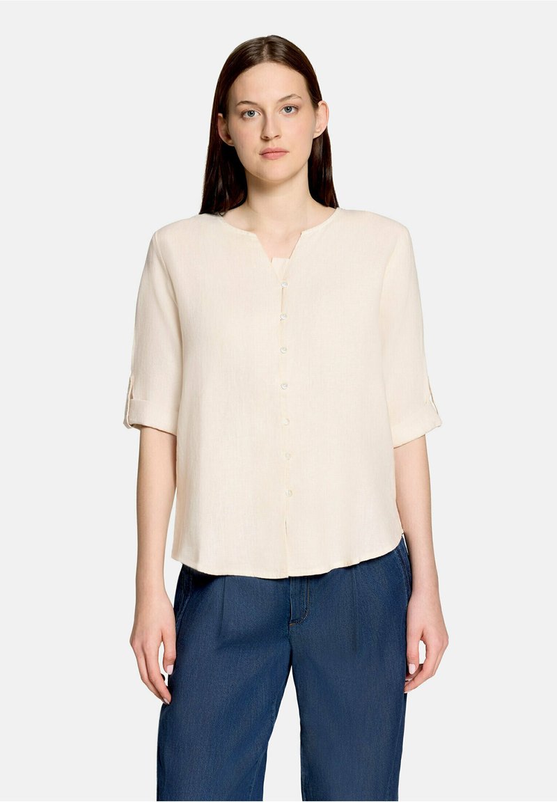 Femme aux cheveux bruns et raides, portant un chemisier crème boutonné aux manches retroussées et un pantalon large bleu, debout devant un fond uni.