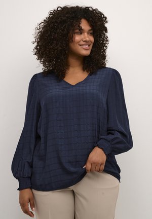 Blouse bleu marine avec un col en V, en tissu texturé, manches bouffantes avec des poignets froncés, et une coupe décontractée. Associée à un pantalon beige clair.