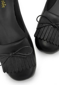 Le ballerine nere in pelle presentano una punta arrotondata, dettagli di frange nella parte anteriore e un sottile fiocco di nastro. La texture è complessivamente liscia.