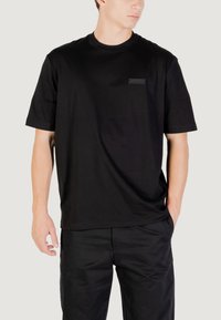 Camiseta de algodón negra con mangas cortas y cuello redondo, presenta una pequeña etiqueta rectangular negra en el pecho y un corte relajado.