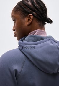 Πλάγια όψη ενός ατόμου με πλεξούδες που φοράει ένα ανοιχτό μοβ φούτερ με κουκούλα, με το κέντημα "HOUDINI" στην πίσω πλευρά της κουκούλας.