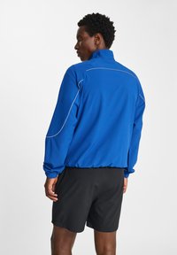 Blå atletisk jacka med hög krage, elastiska ärmmuddar och kontrasterande vit sömnad, kombinerad med svarta shorts. Slät tyg, sportig design.