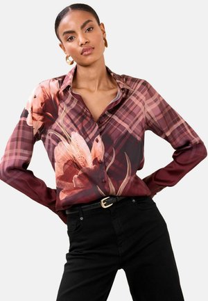 Kukka-kuvioinen burgundinpunainen ja vaaleanpunainen silkkipaita, jossa on kaulus, pitkät hihat ja napitus, yhdistettynä mustiin korkeavyötäröisiin housuihin.