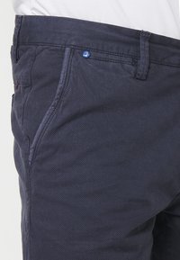 Close-up van marineblauwe getextureerde broek met zijzak en ceintuurlus, gedragen met een wit shirt, met details van de stof en stiksels.