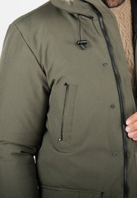Parka d'hiver vert olive avec fermeture éclair à l'avant, boutons-pression, accents de fermeture éclair noirs, capuche avec cordon de serrage et poche latérale avec rabat.
