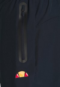 Marina blå tygbyxor har en sidodragkedjeficka med en svart panel och en flerfärgad logopatch nederst.