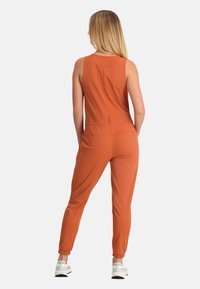 Salopette arancione senza maniche con gambe affusolate, texture liscia, polsini elastici e dettagli sulla vita posteriore. Il modello indossa sneakers bianche.