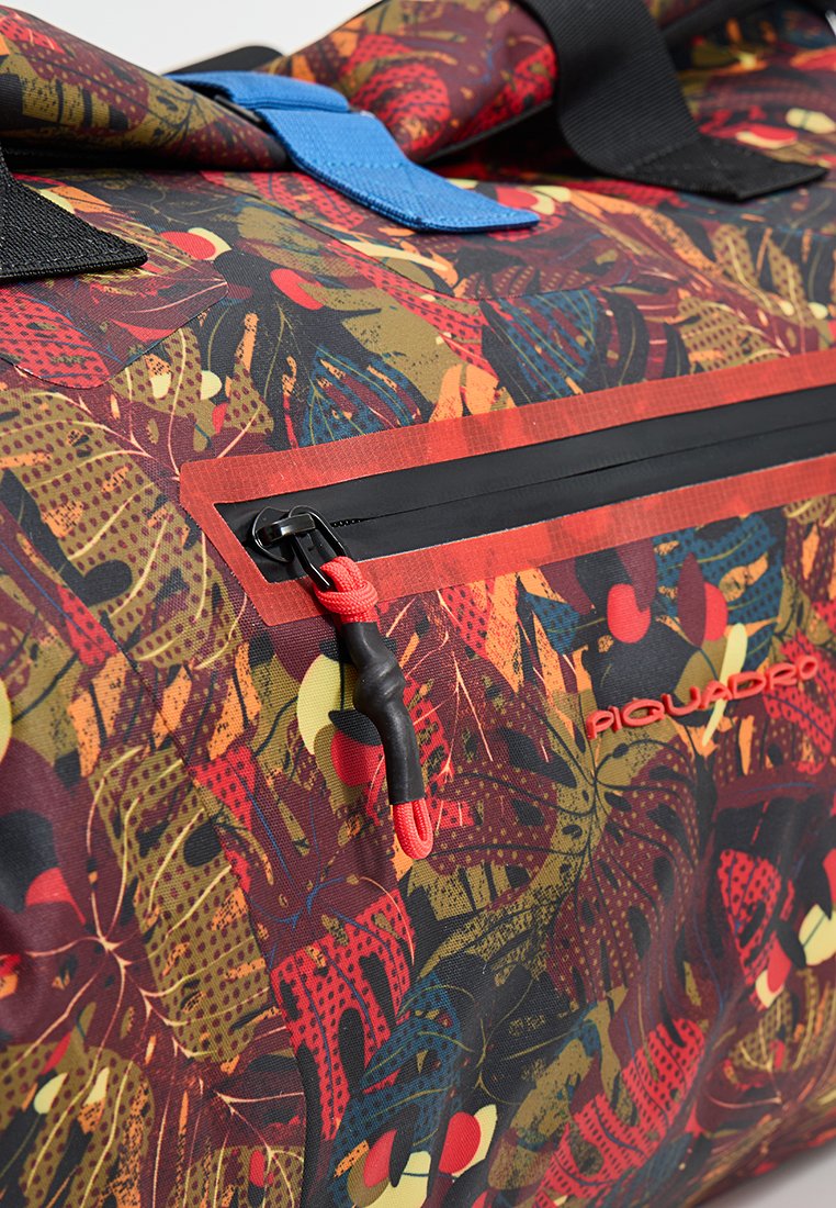 Borsa con motivo astratto di foglie colorate, cerniera nera con anello rosso, tracolla blu e logo "Piquadro" rosso su tessuto.