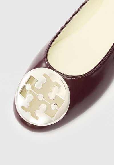 Bordová kožená balerína s okrúhlym kovovým logom na špičke, hladkým povrchom a ľahkým dizajnom.