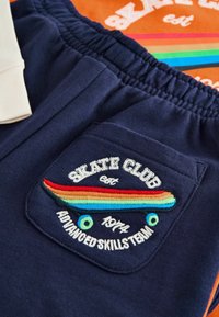 Marineblaue Trainingshosen mit einem Patch, der ein mehrfarbiges Skateboard-Design und den Text "SKATE CLUB" zeigt. Elastischer Bund und weicher Stoff.