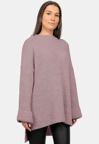 Vrouw draagt een loszittende, gestructureerde mauve trui met lange mouwen en zwarte legging, staande tegen een effen lichte achtergrond.