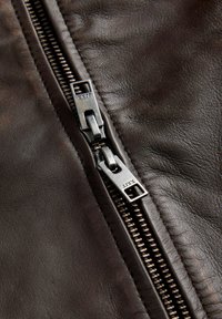 Detailný pohľad na dva kovové posúvače zipsu na tmavohnedom koženom odevu, zobrazujúce textúru a detaily šitia.