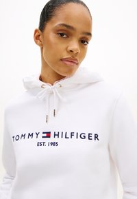 Biała bluza z kapturem z regulowanym sznurkiem i złotymi elementami, z niebiesko-czerwonym haftowanym logo "TOMMY HILFIGER EST. 1985" na przedzie.
