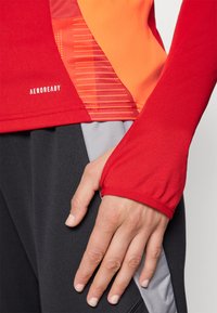Rotes sportliches Langarmshirt mit orangen Akzenten und einem strukturierten Seiteneinsatz, mit enganliegenden Bündchen. Logo an der Seite sichtbar.