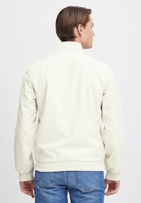 Lys beige, vandafvisende jakke med høj krave, ribbede manchetter og kant. Har en glat, let blank overflade.