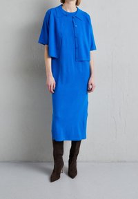 Blå stickad klänning med korta ärmar, knäppt fram och en klassisk krage, kombinerad med höga bruna mockastövlar. Slät textur.