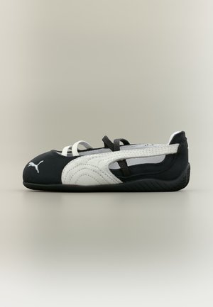 Chaussure plate de ballet Puma noire et blanche avec des sangles élastiques blanches et un logo sur l'orteil, vue de côté sur fond uni.