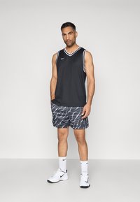 Μαύρο αμάνικο μπλουζάκι με λευκή προβαλλόμενη, συνδυασμένο με μαύρο σορτσάκι που διαθέτει κυματιστό λευκό μοτίβο. Φορεμένο με λευκά αθλητικά παπούτσια και κάλτσες.