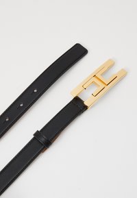 Black leather belt with multiple holes and a gold rectangular buckle featuring a cut-out geometric design, laid flat on a white surface.