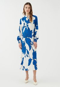 Langes, weißes Kleid mit gro�ßem blauen Blumenmuster, hohem Halsauschnitt und langen, puffärmeligen Ärmeln. Verfügt über eine Knopfleiste und gefaltete Akzente.