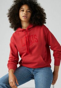 Felpa rossa in cotone con tasca a marsupio, cappuccio con cordino e logo in rilievo. Abbinata a jeans di denim blu, per un look casual.