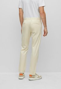 Pantaloni color crema con una vestibilità slim, caratterizzati da un design con tasca posteriore e cuciture sottili. Abbinati a sneakers beige con accenti arancioni.