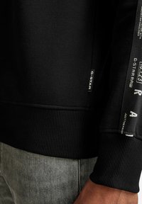 Sudadera negra con dobladillo y puños acanalados. Presenta acentos de marca en la manga y una etiqueta lateral. La tela parece lisa y gruesa.
