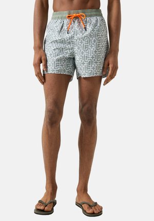 Man draagt een patroon zwemshorts met groene tailleband en oranje trekkoord, staat blootsvoets in slippers tegen een witte achtergrond.