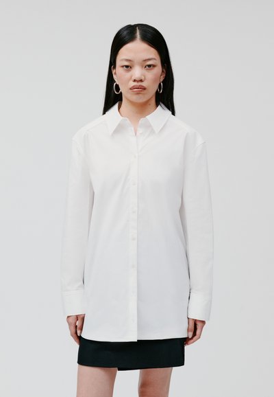 Camisa blanca de cuello con botones en la parte delantera, de mangas largas y corte relajado. Material de algodón suave, combinada con una falda corta negra.